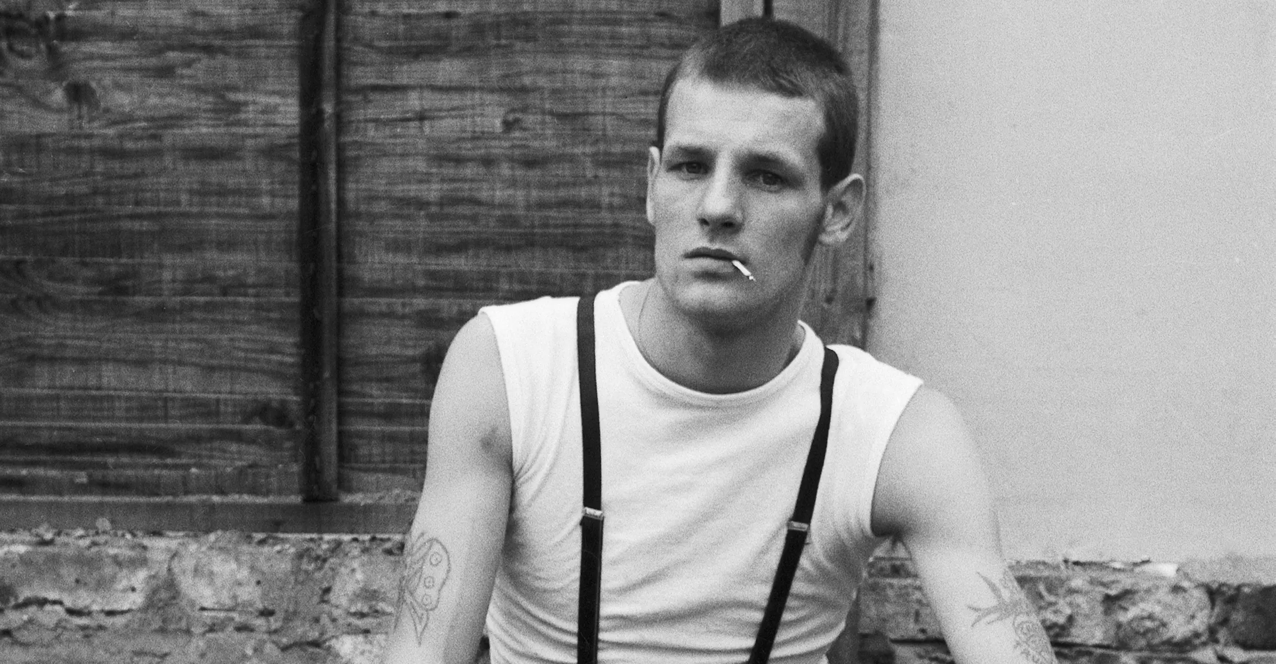 Black and white photograph of a young man in a white sleeveless vest and braces, short cropped hair and cigarette dangling from mouth, staring into at the camera.