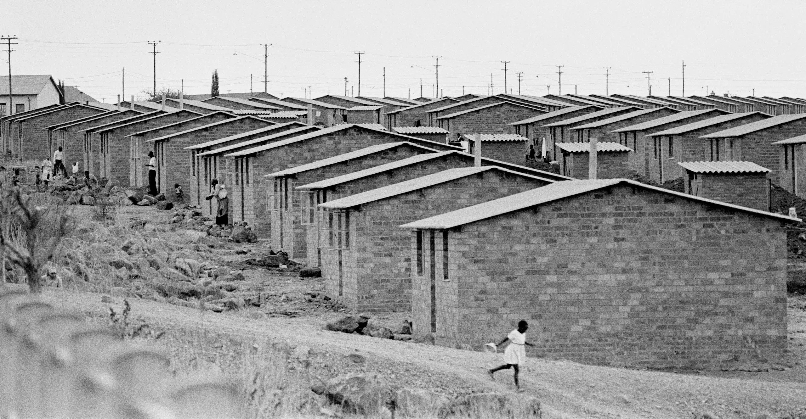 Black and white landscape photograph of a row of houses. A child can be seen walking along a pathway and a group of people are stood outside the houses in the distance.