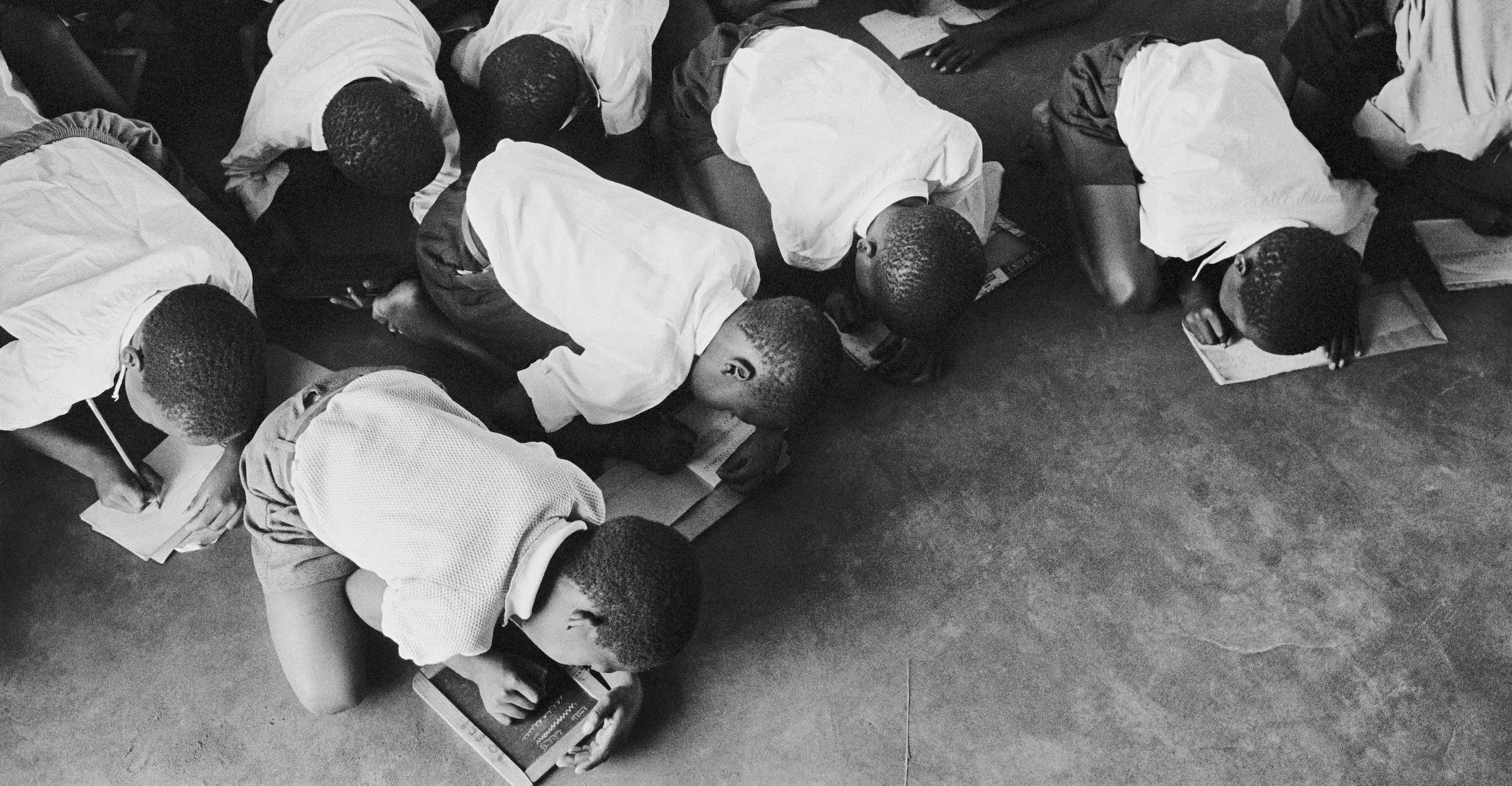 Black and white photograph of several schoolchildren bent over on the floor to write, some write on paper others write on chalkboards.