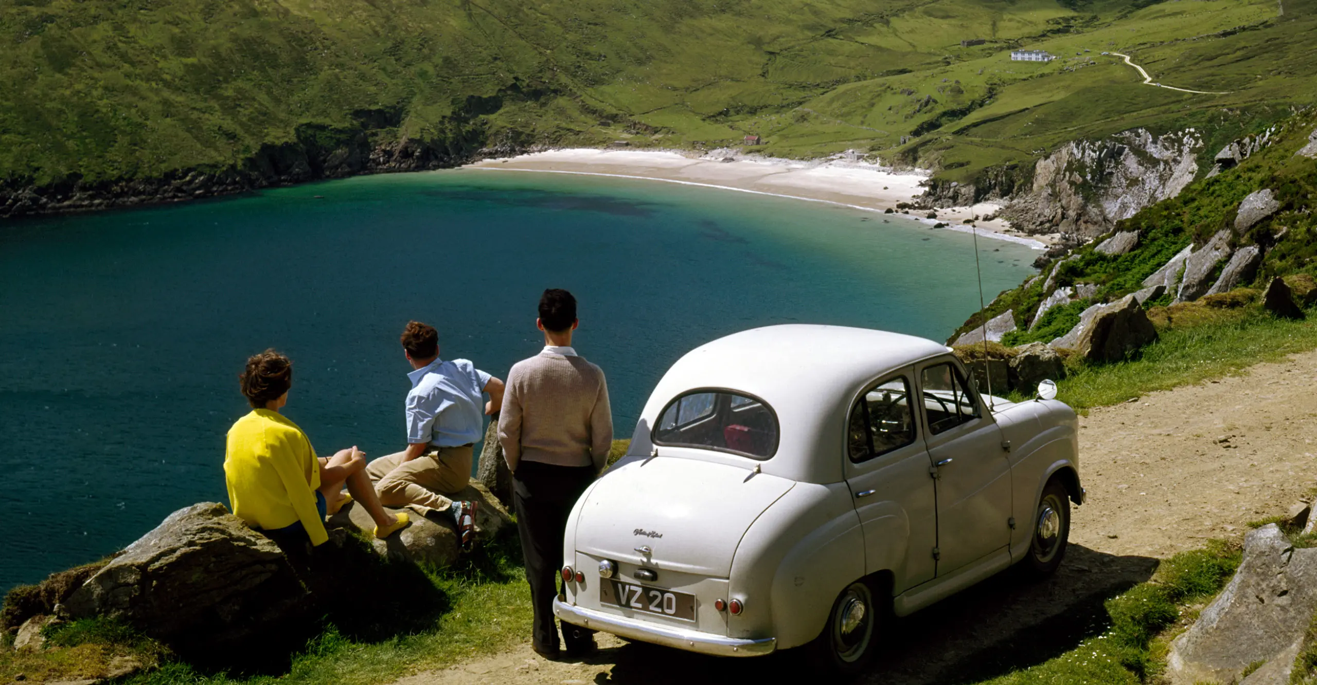 High-saturated image of three people in late 40s casual clothingstanding outside their white car overlooking out to a bay on a sunny day
