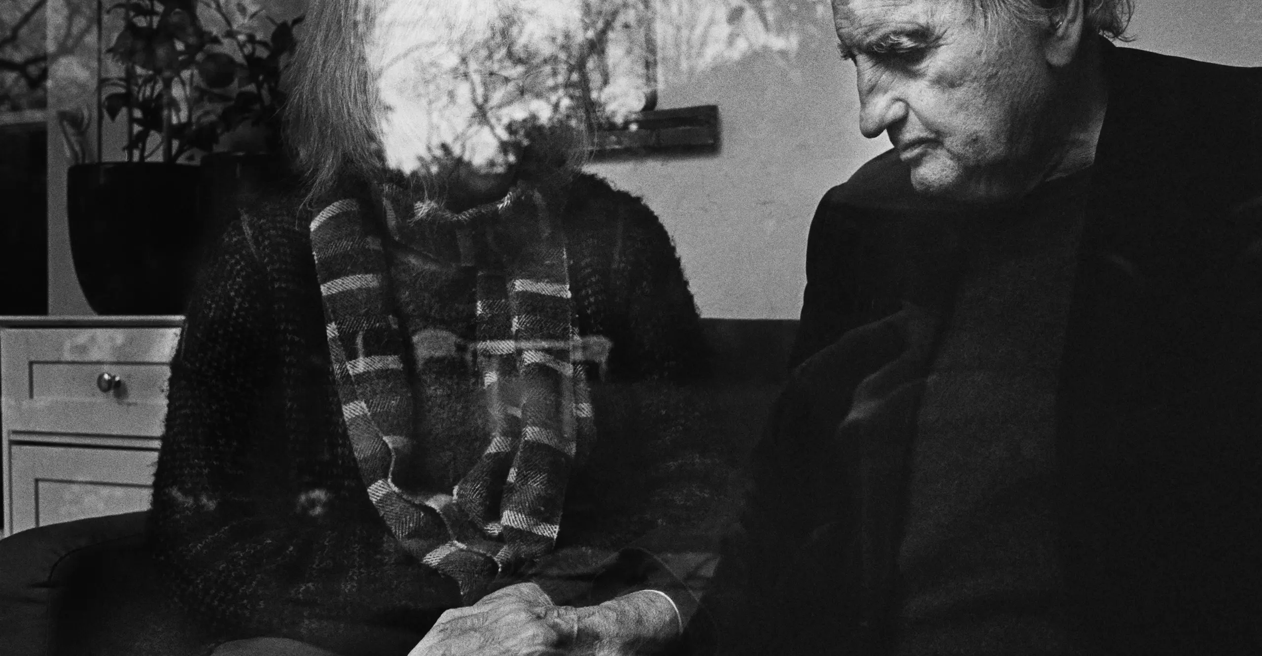 Black and white image featuring an older couple with the person on the right's face partially obscured by a reflection on glass. Man's hand is on the other person's knee.