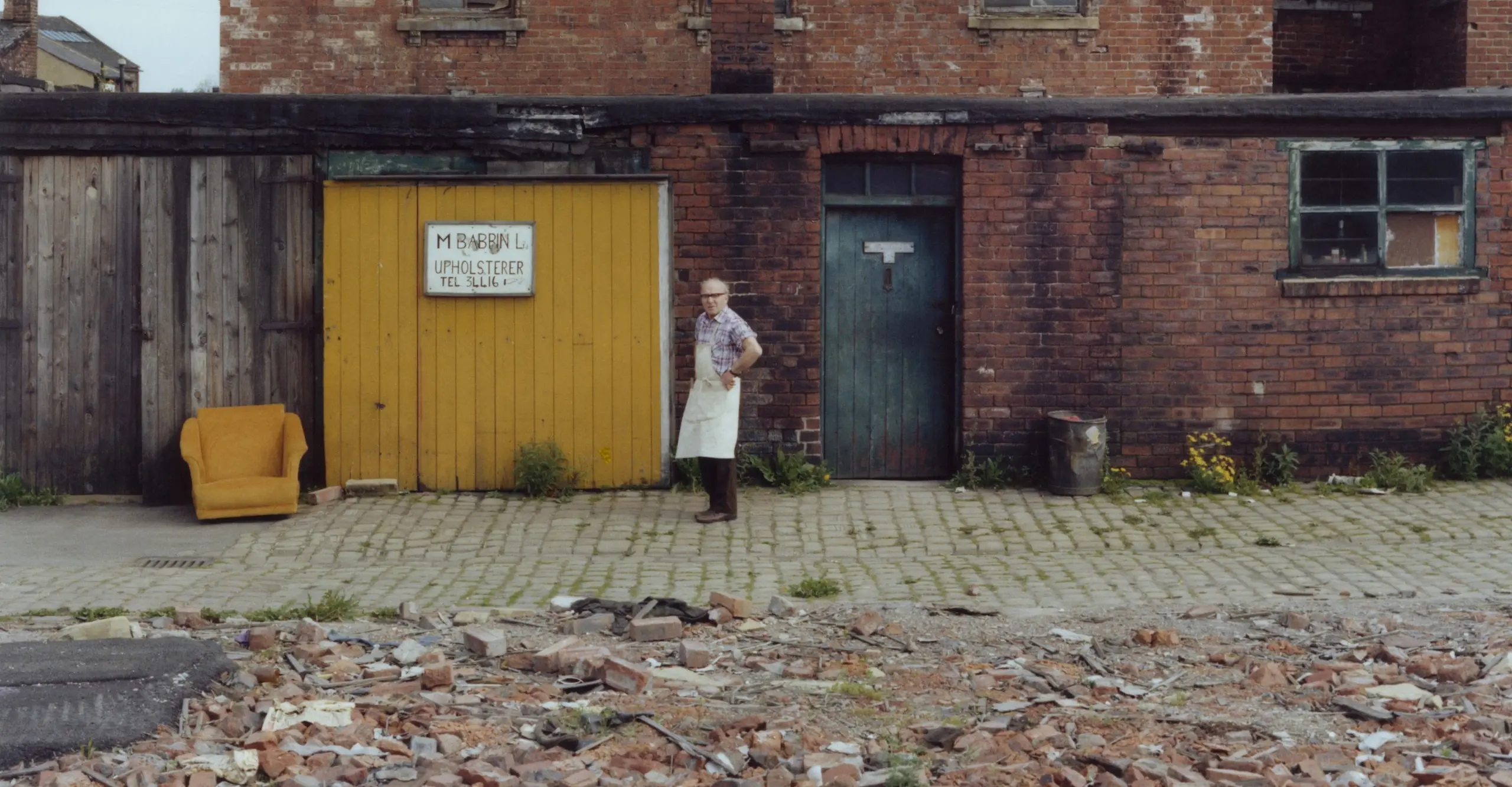 Colour photograph of a man in an apron standing on the street, outside of his upholstry shop.