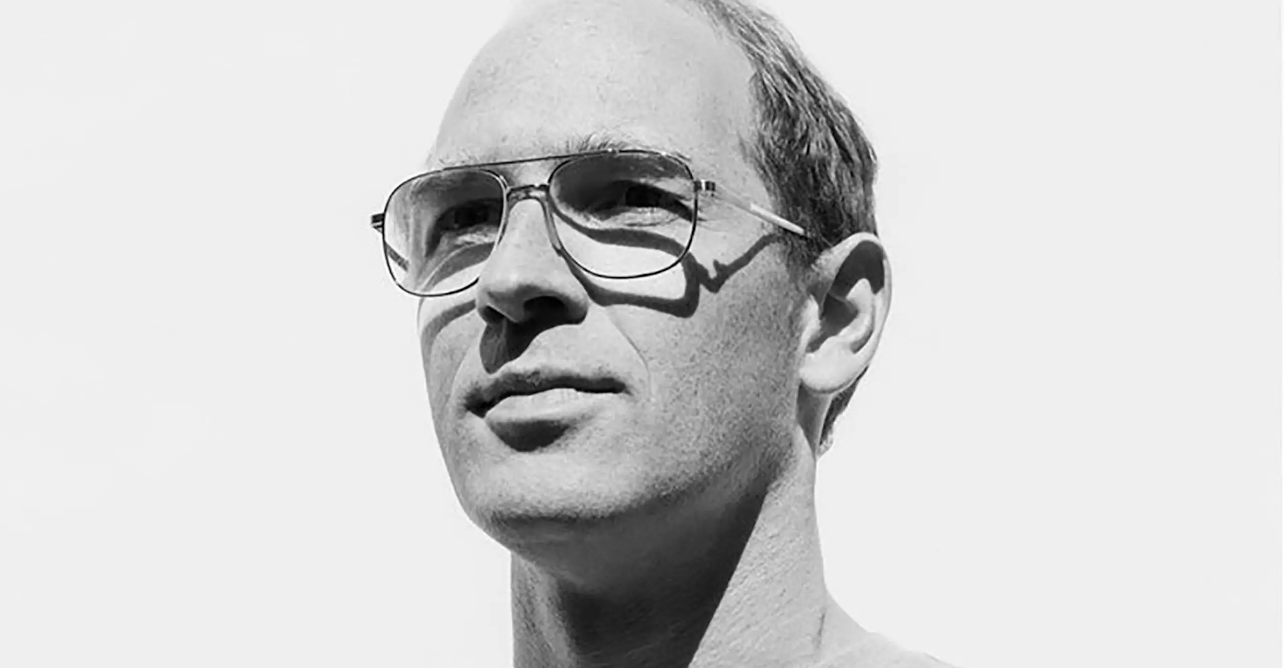 Black and white head and shoulders portrait of a white man wearing aviator style glasses looking off to the right smiling slightly.