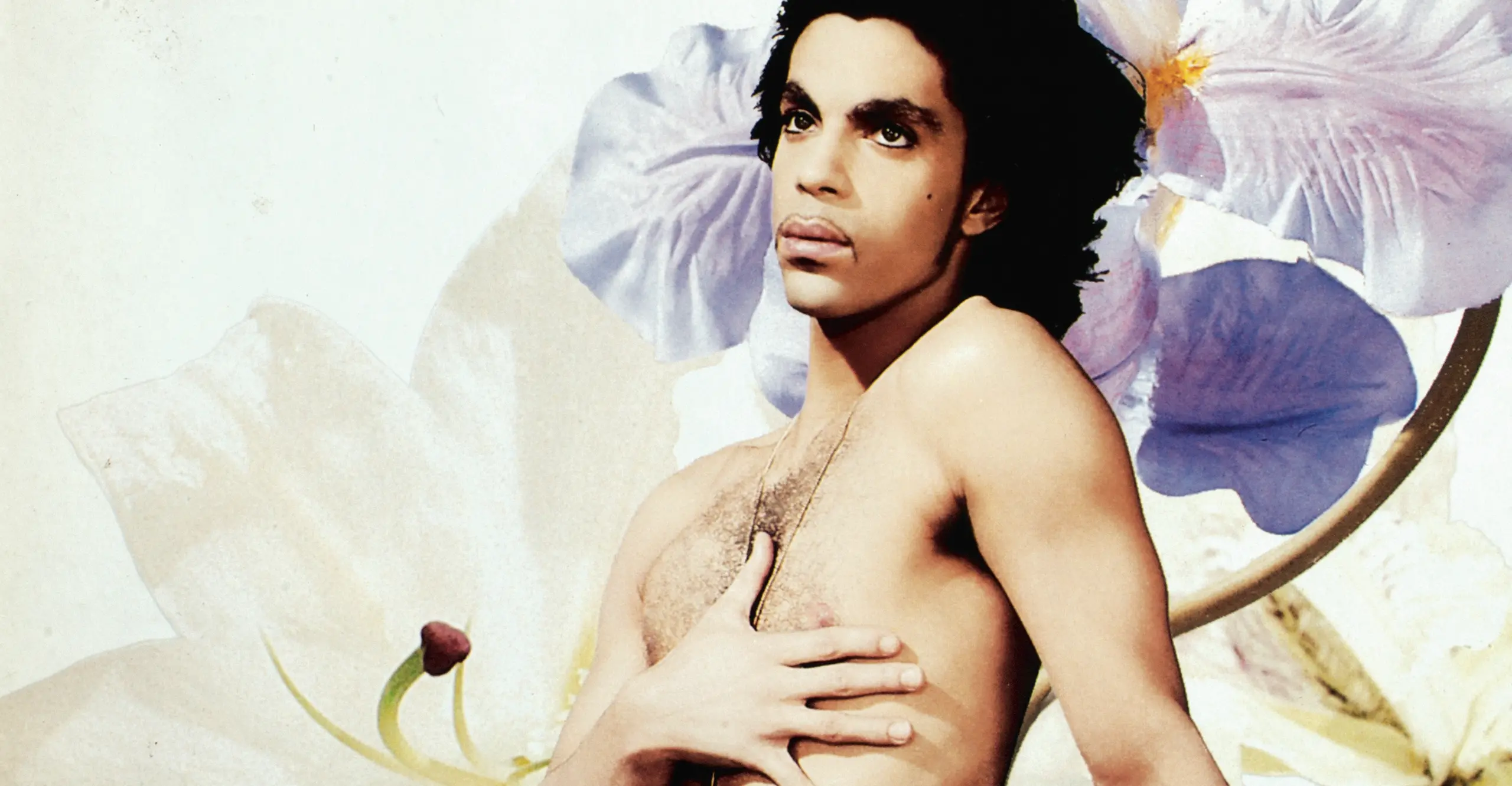 The musician Prince poses naked, eyes looking dolefully upward, against a large flower backdrop.. 