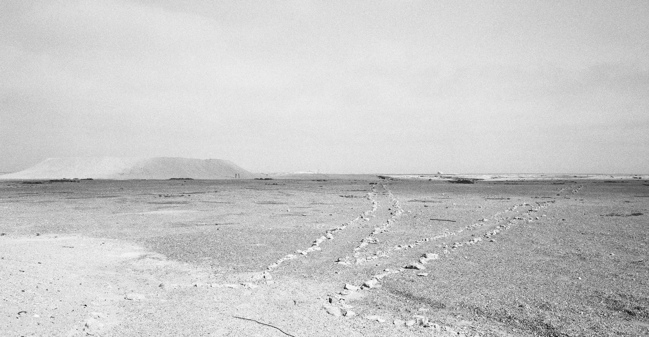 Black and white photograph of a desert landscape with two stone line paths leading off into the distance where a large crater and smaller buildings sit on the horizon.