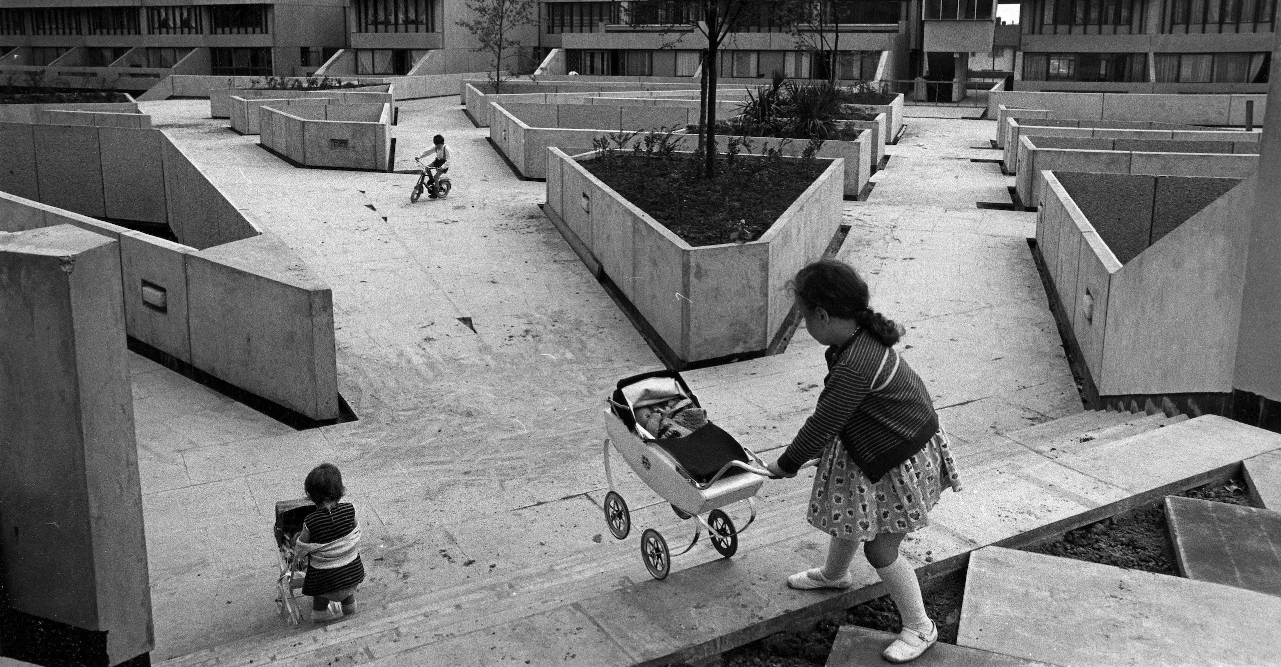 A black and white photo showing two children pushing prams on stairs in a concrete courtyard while another cycles in the distance.