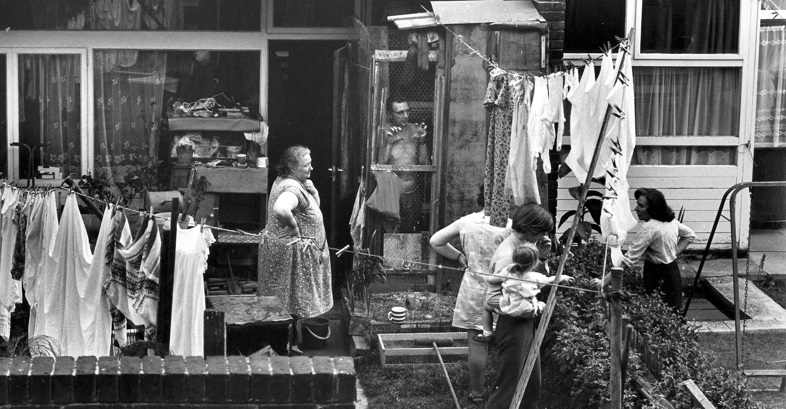 Black and white photograph of families talking over their shared low garden wall with laundry hanging.