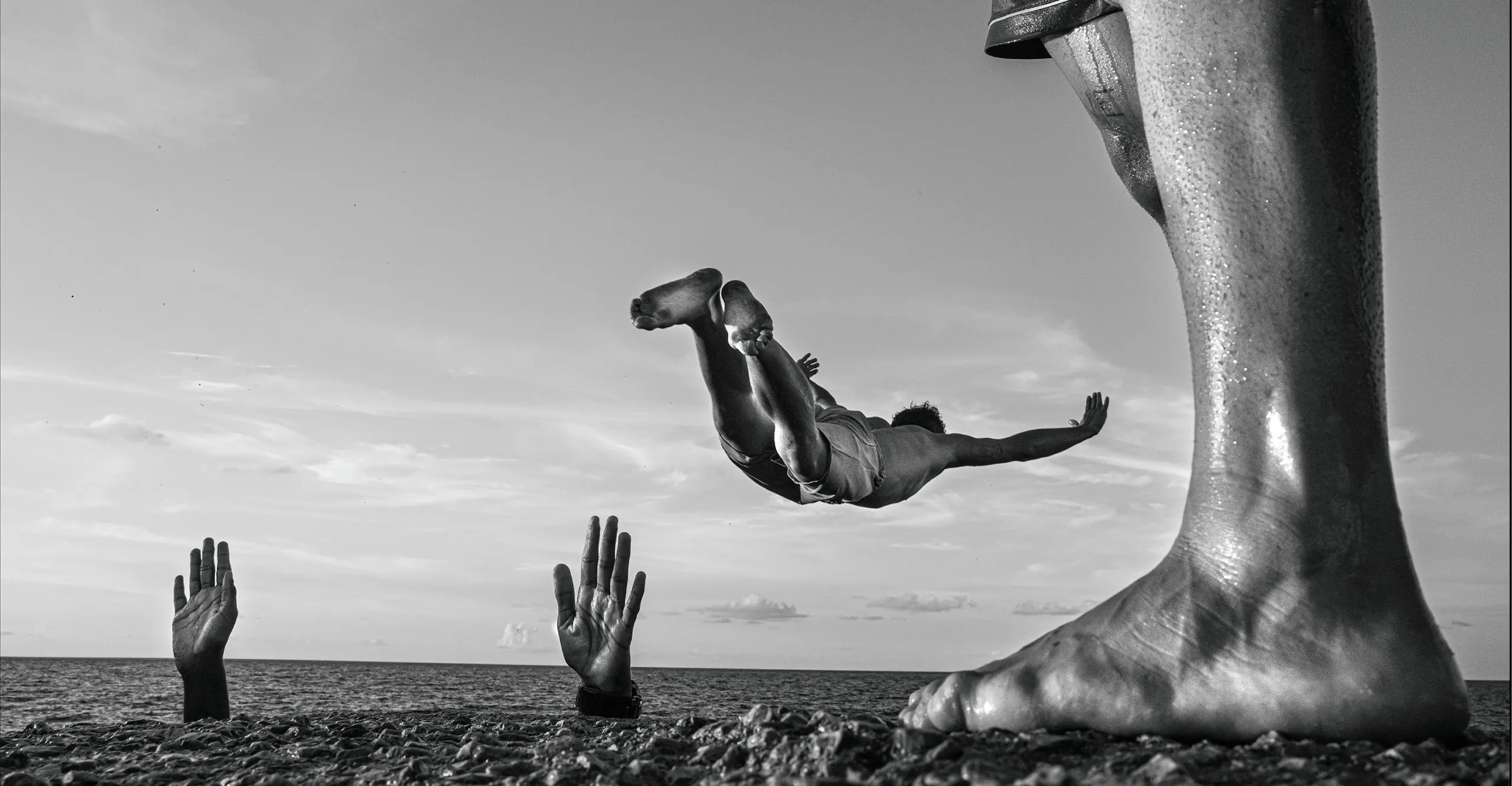 Black and white photograph of a beach scene with a person's leg in the foreground and a person mid air about to dive into the water in the background