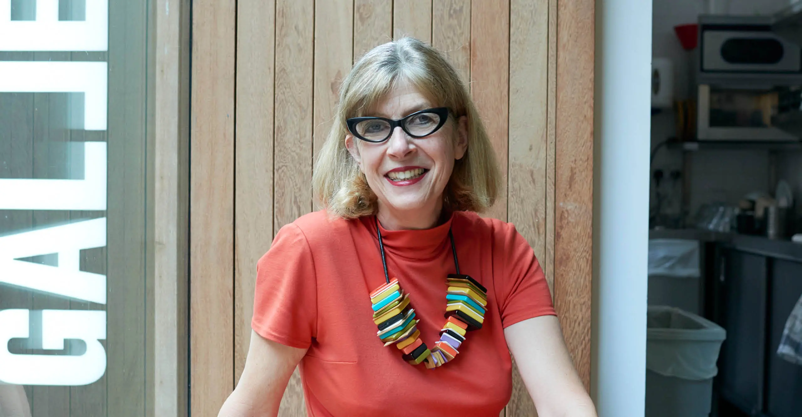Portrait of a woman with short hair, bright coloured orange dressed an a multi-coloured necklace, sitting down inside The Photographers' Gallery
