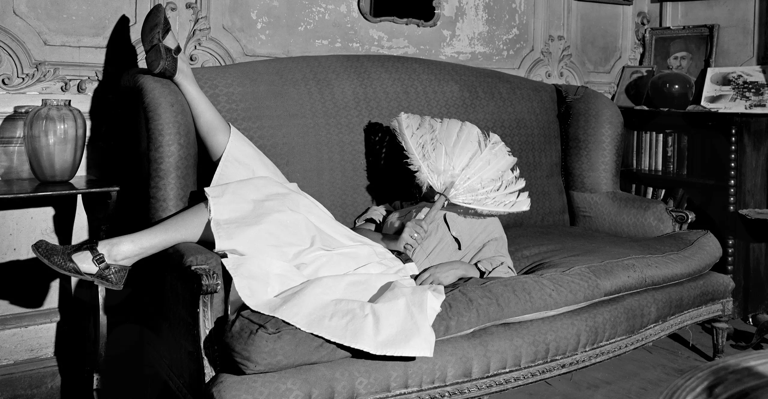 Black and white photograph of a woman laying down on a sofa, holding a feather fan over her face 