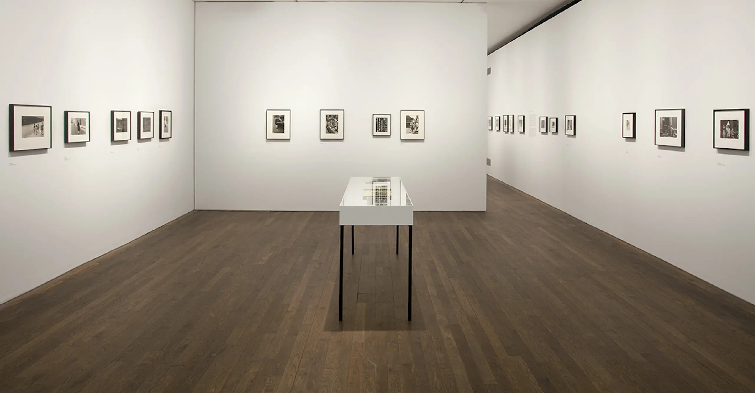A white gallery space with small black and white prints on white walls and a vitrine in the centre.