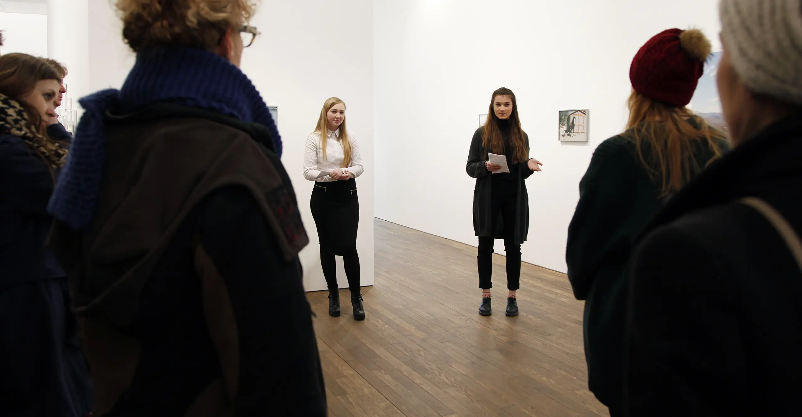 A photograph of a person in the middle of the frame pointing to a photograph on a gallery wall.