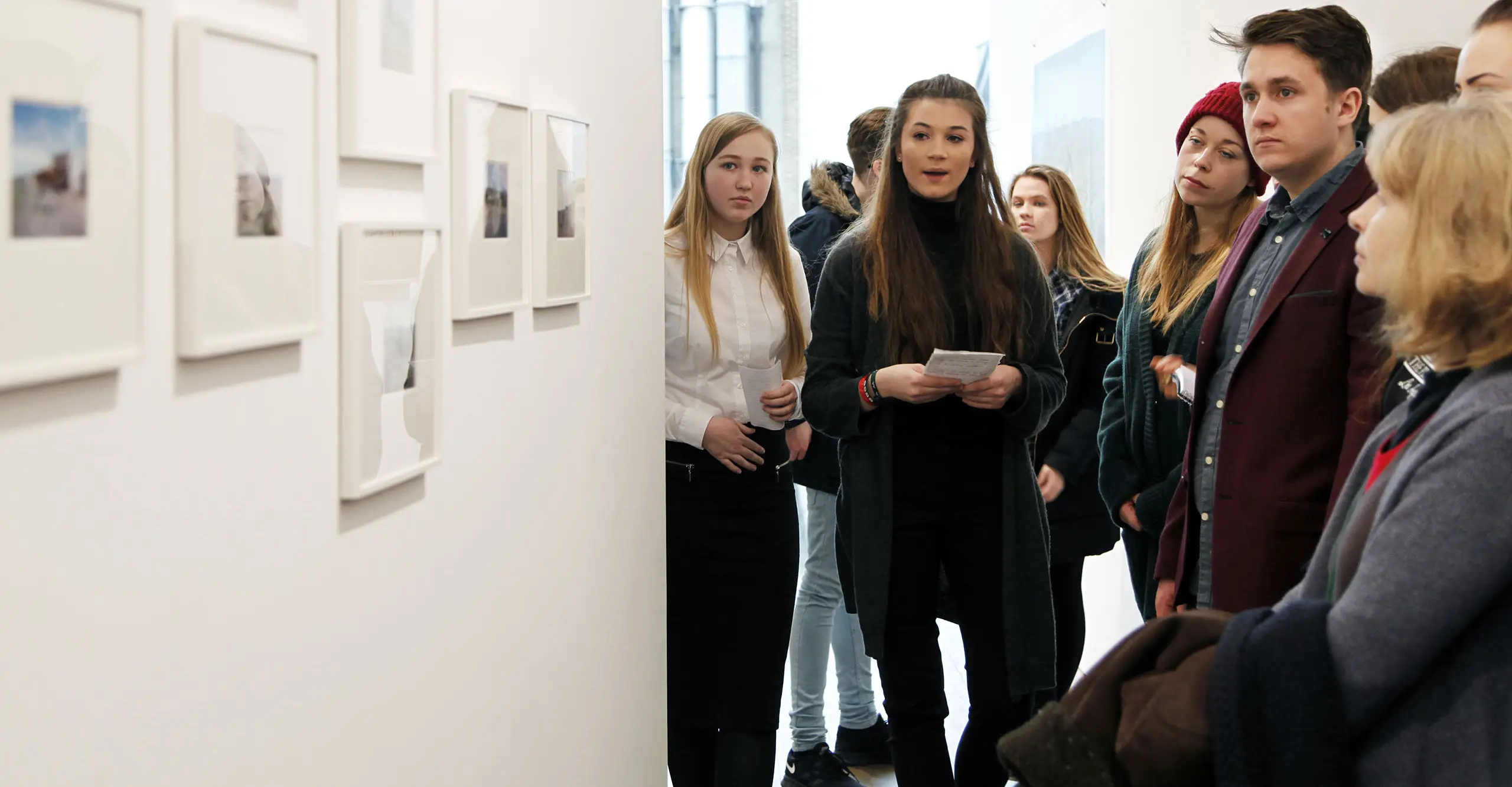 A photograph of a young person speaking to a group of people in a gallery.