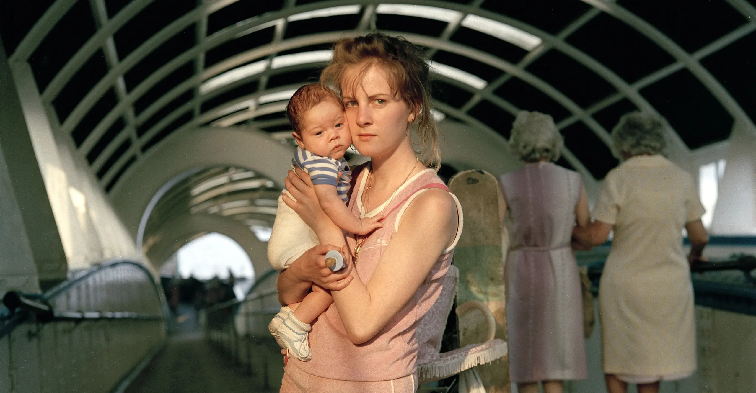 A young woman in a pink tracksuit looks directly at the camera, holding a baby