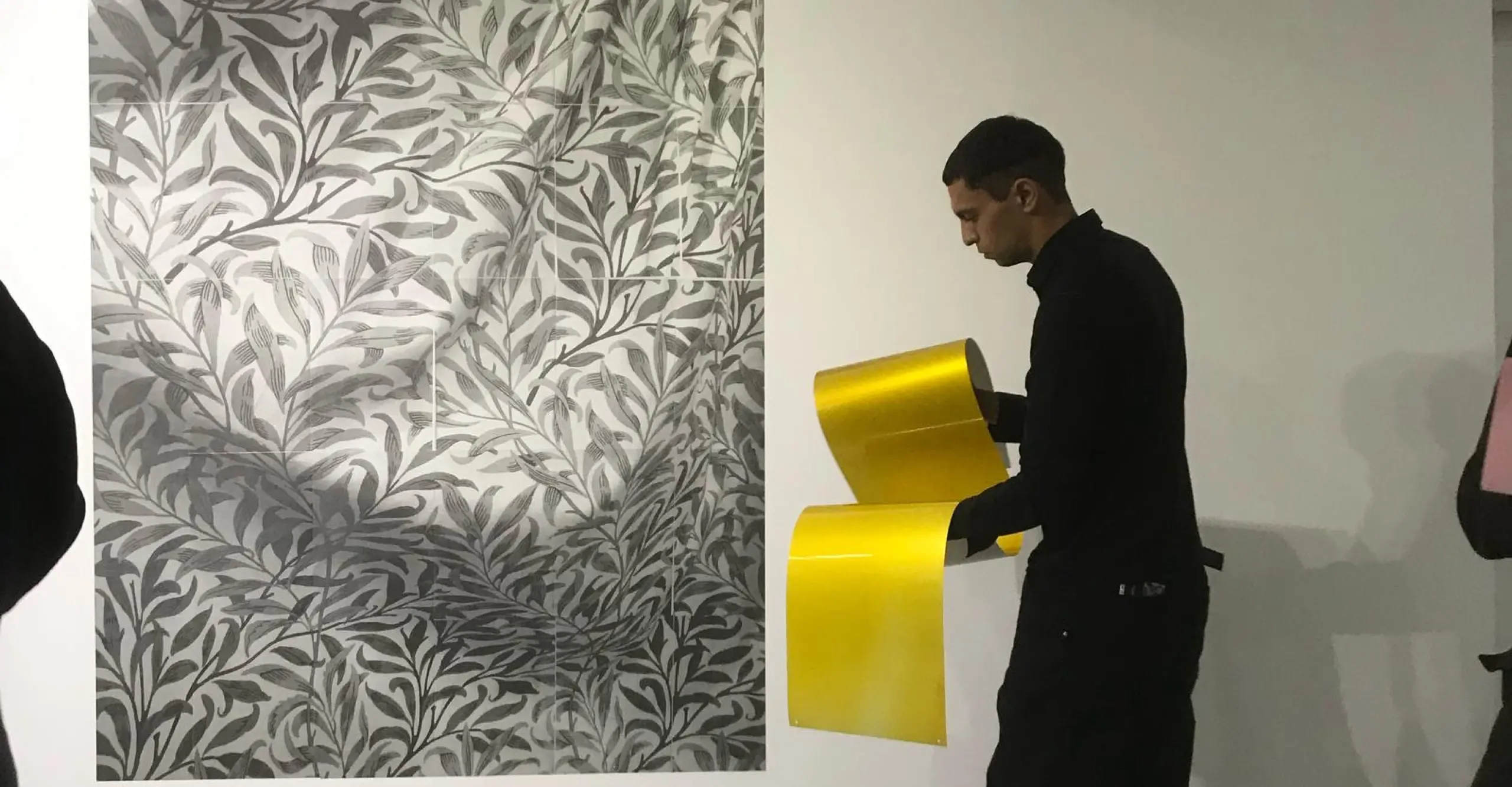 Figure in all black holding gold paper in front of an abstract, grayscale work in a white room