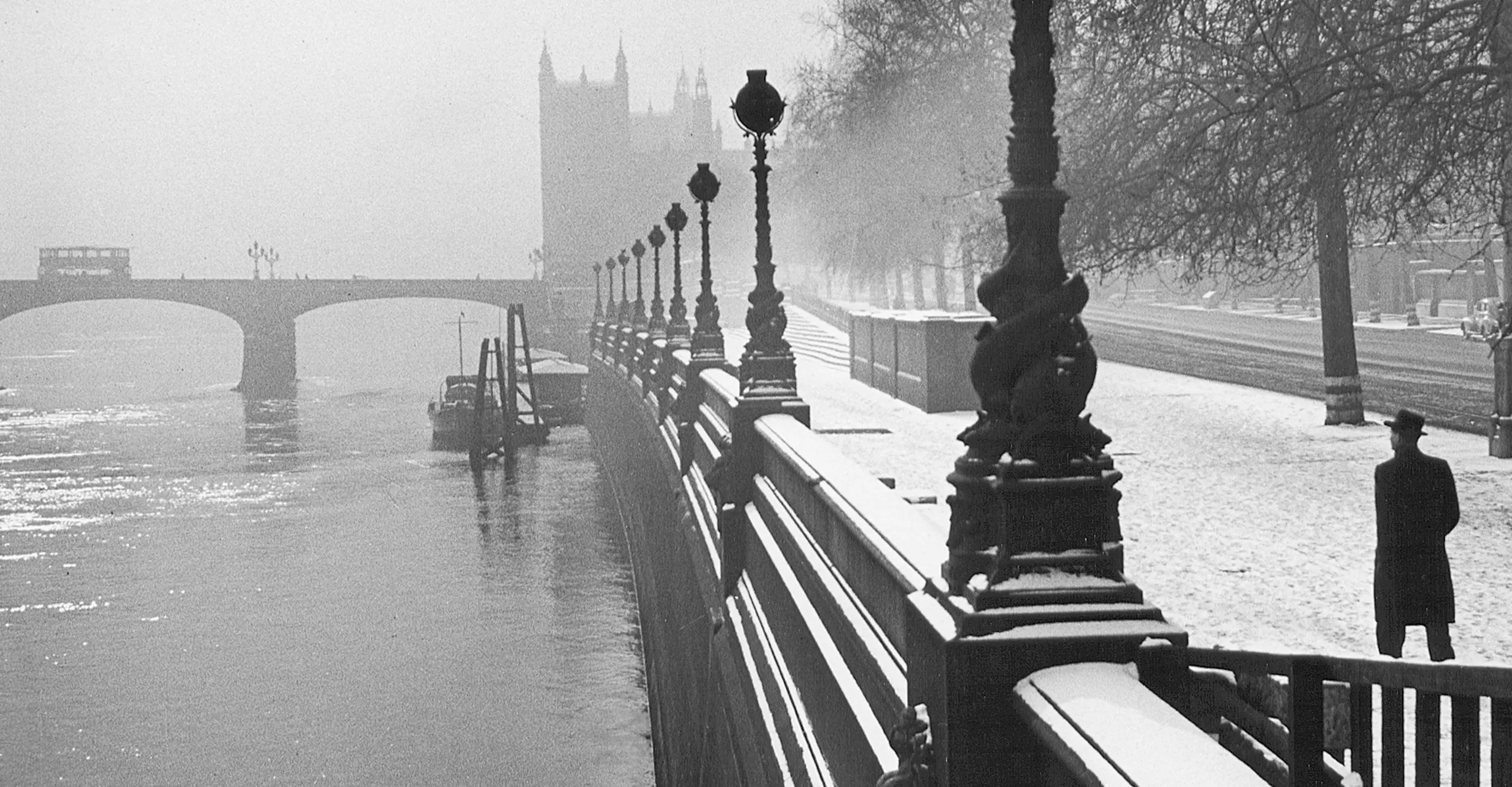 Black & white snowy scene of London's Embankment in the 40s with a man in a hat walking along the bank