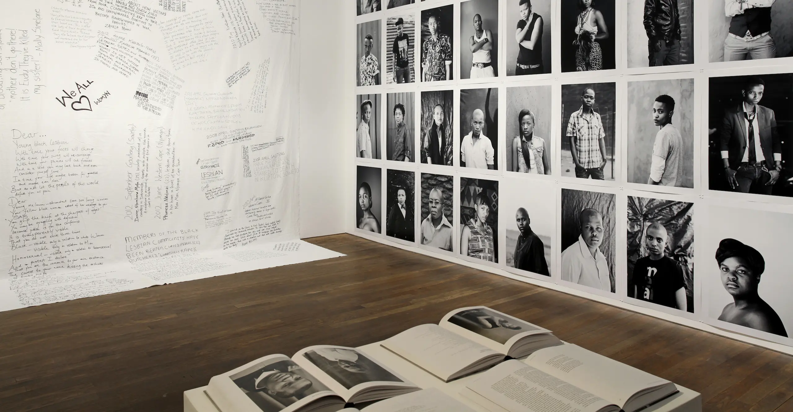 The corner of an exhibition space with a photography book on a plinth in the foreground, black and white portrait photographs along one wall and handwriting on a white background along the other wall. 