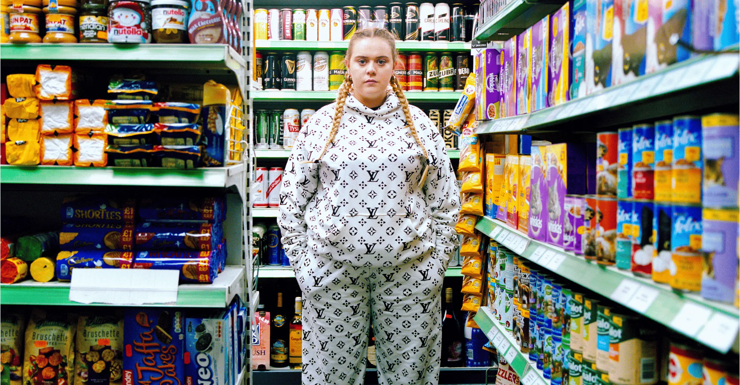 A colour image of a young woman, wearing her blonde hair tied into a braid and a white tracksuit on, standing in the middle the aisle of a well stocked grocery shop aisle. 
