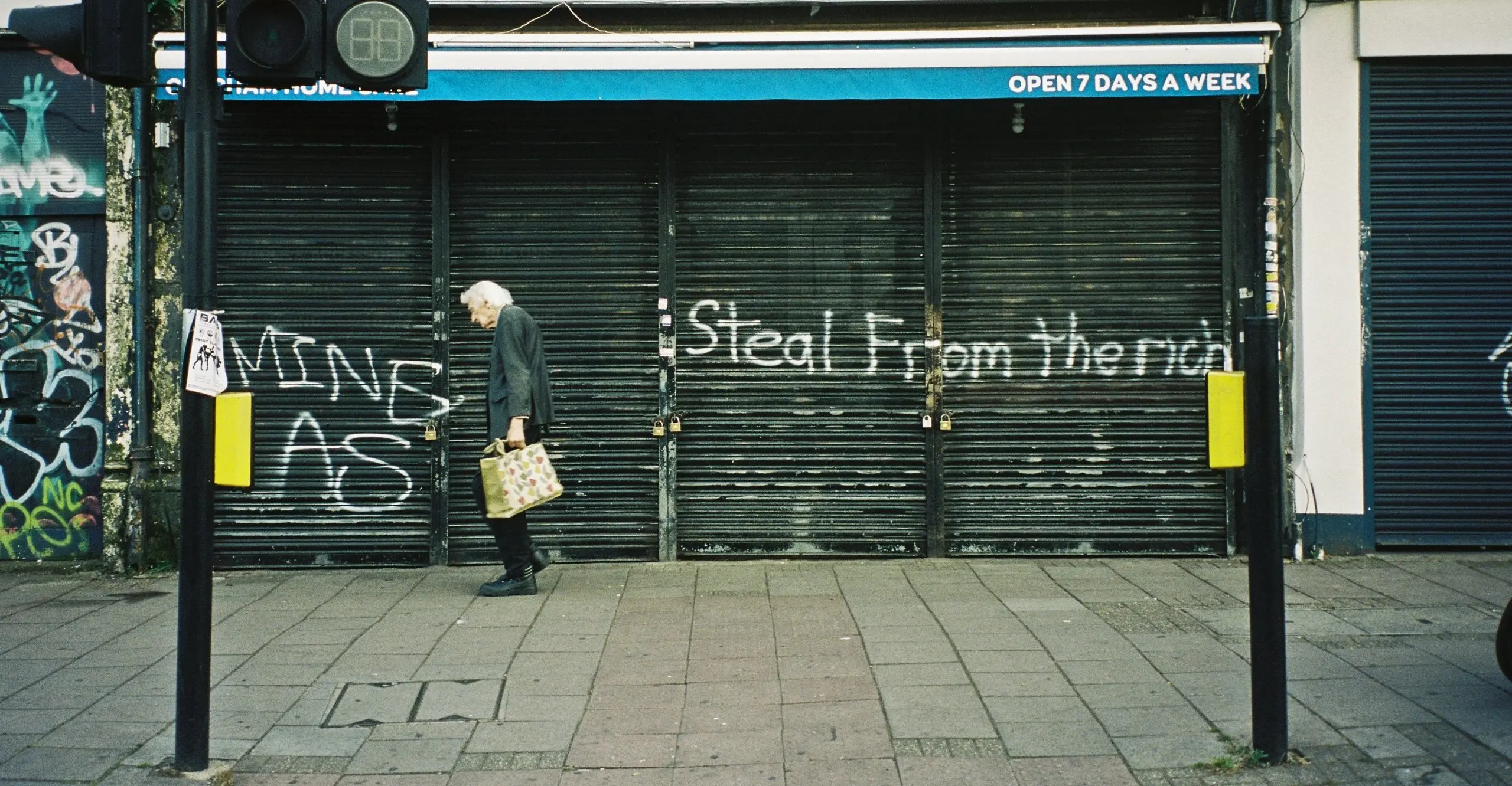 Colour photograph of an elderly person walking along the street past a shuttered shop front that has “STEAL FROM THE RICH” spray-painted in front.