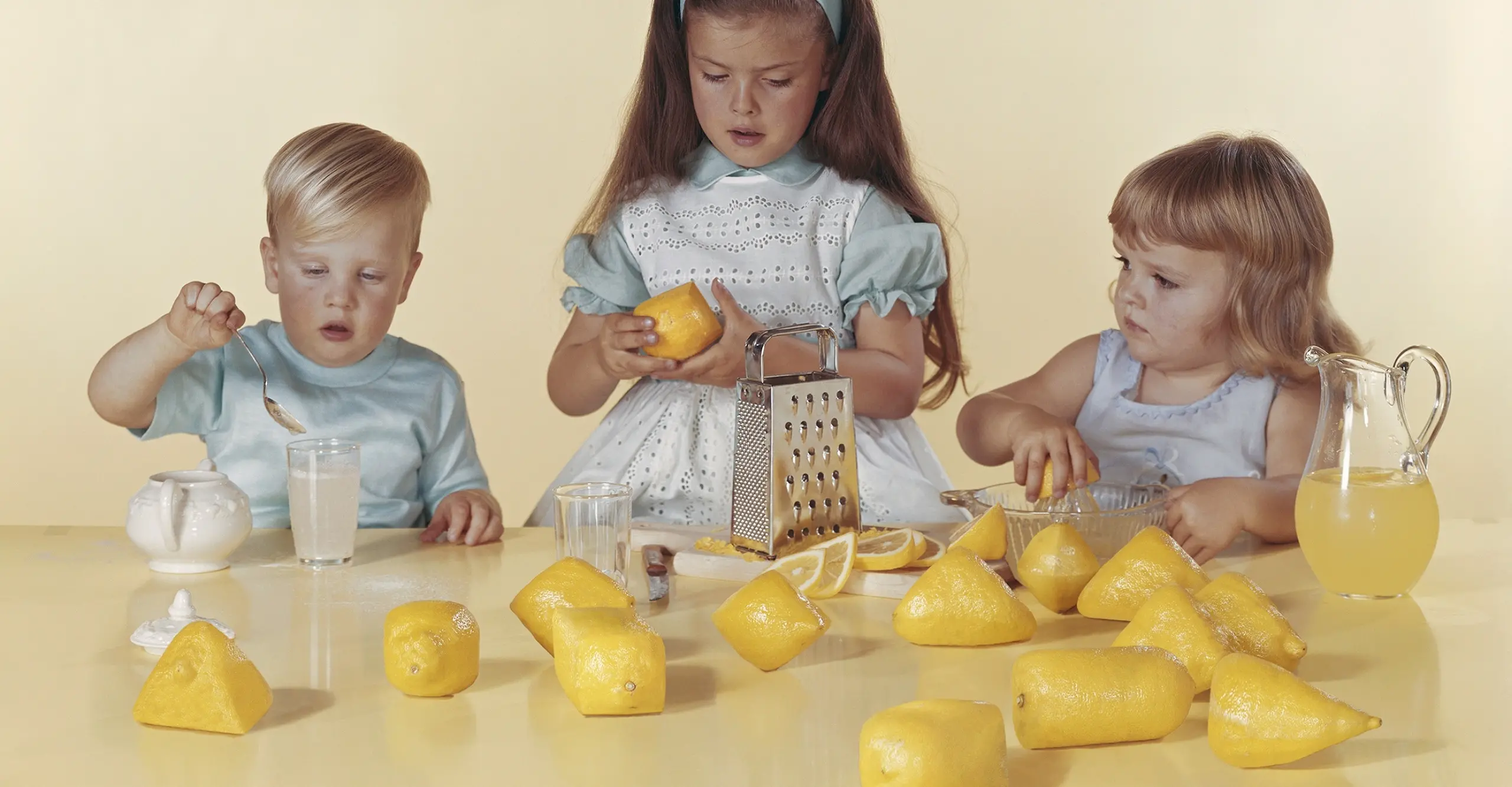 Colour photograph of three children sat at a table with lemons spread out, along with a grater and a jug.