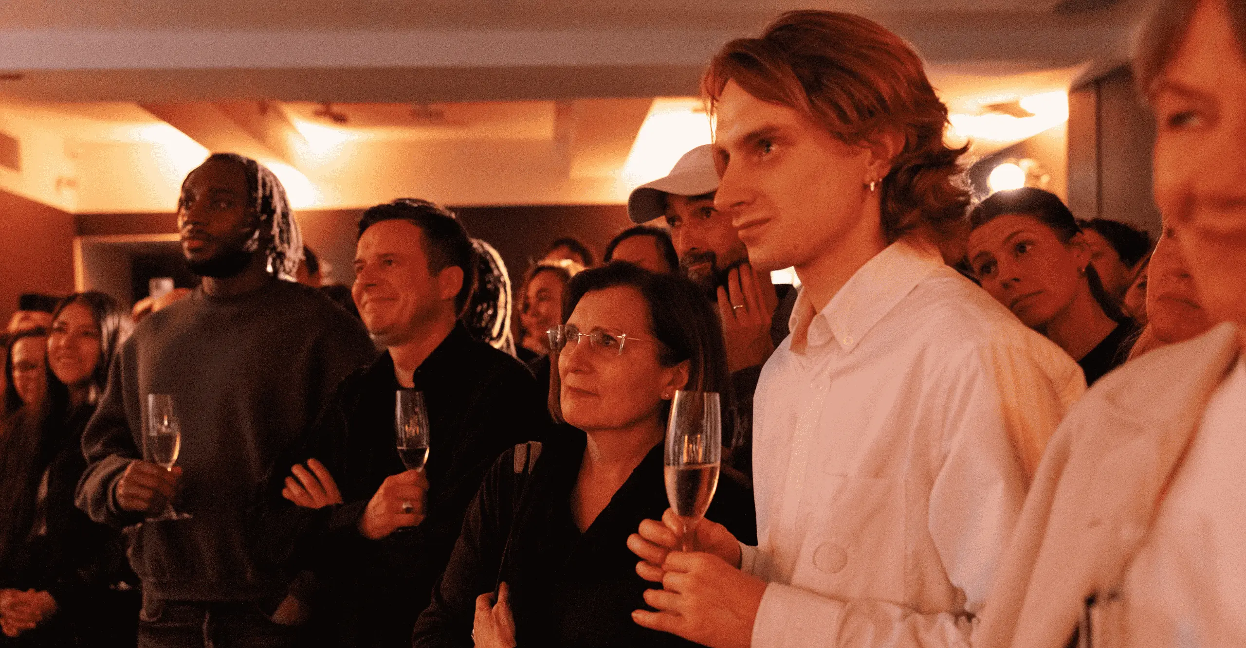 People are stood in a small crowd at an event looking over at something out of shot. The lighting is warm toned and they have glasses of champagne in their hands.
