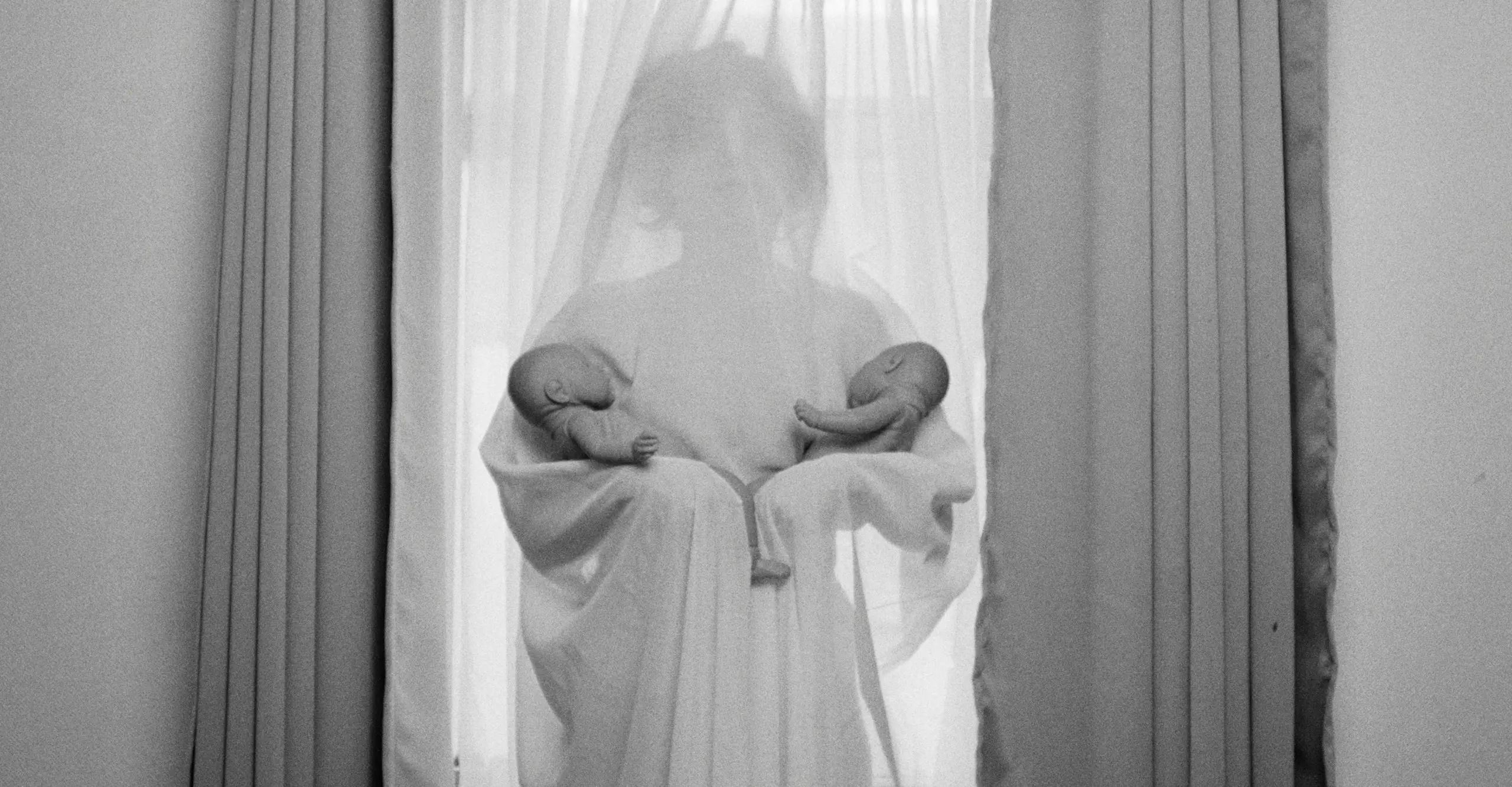 Black and white photograph of a woman behind a net curtain, holding two babies in each of her arms.