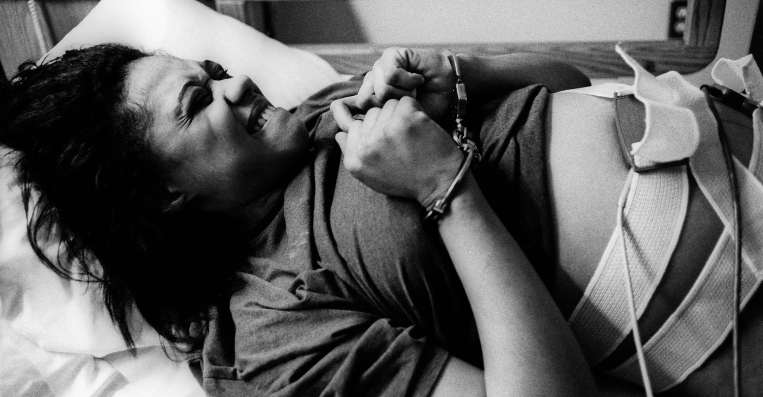 Black and white photograph of a woman handcuffed in a bed with tubes and straps taped to her stomach.