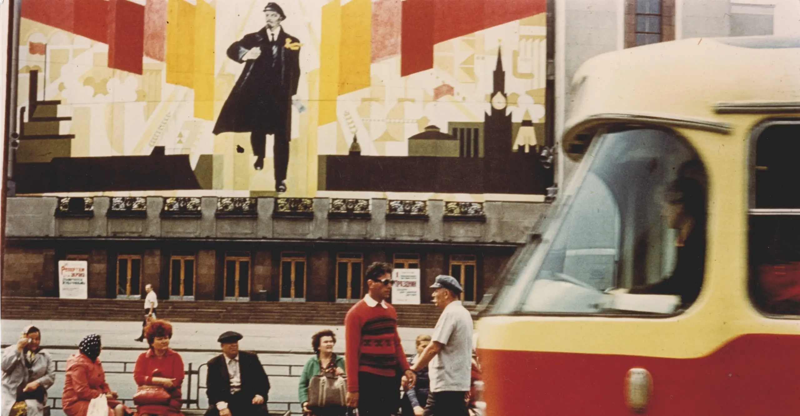 Colour photograph of people sat on a bench  with a large billboard of Vladimir Lenin behind. A tram can be  seen in the corner passing in front.