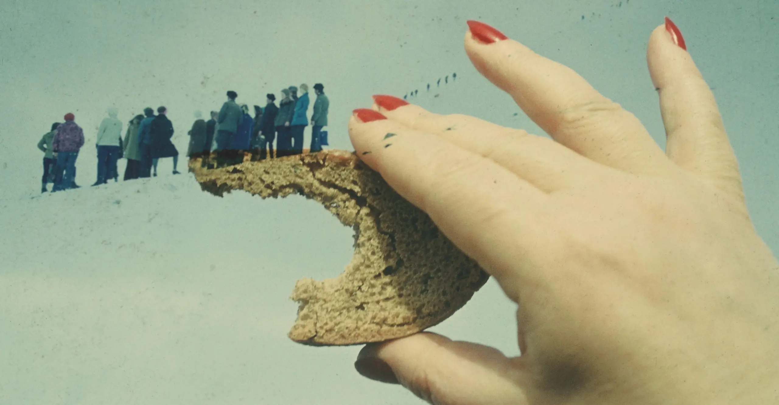 A vintage double exposed image of a hand with painted red nails, holding a crust of bread. An overlay of a group of people in winter coats are seemingly walking down the top of the crust of the bread.