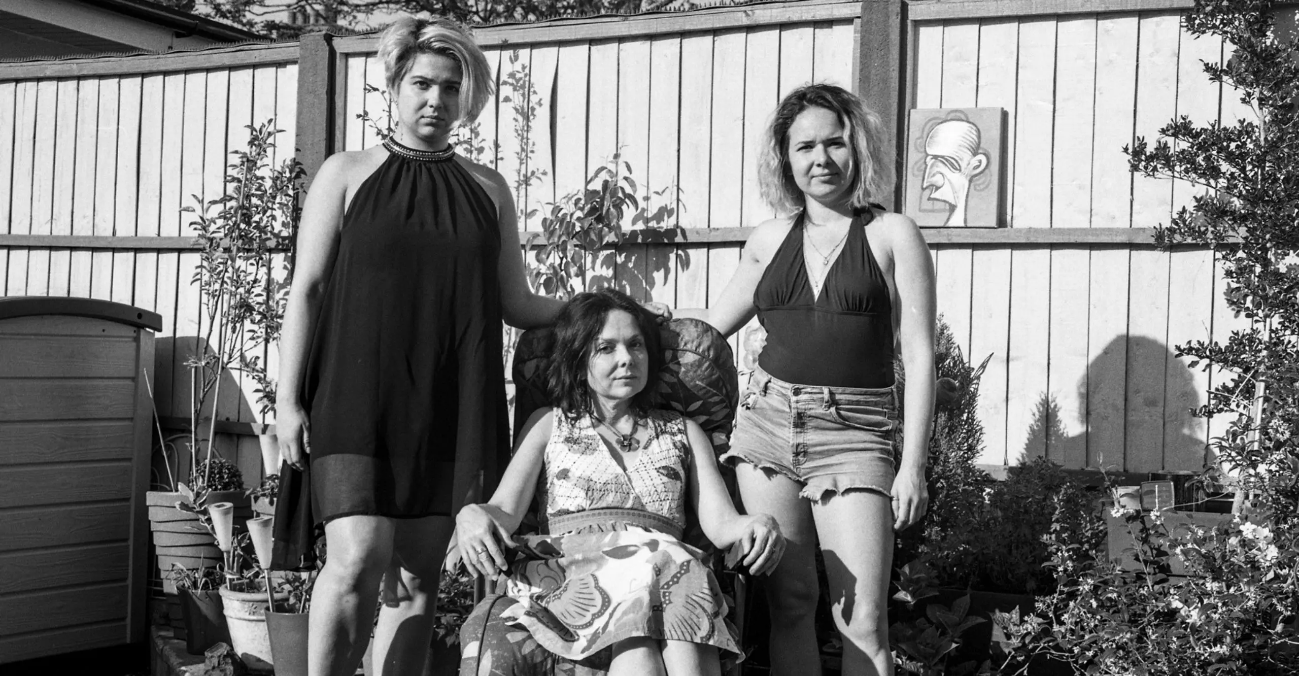 A black and white photo of three women in the same family of different generations sat for a portrait in a driveway. The youngest sits on an outdoor garden chair whilst the two elder women stand behind her by each shoulder. They look content and relaxed.