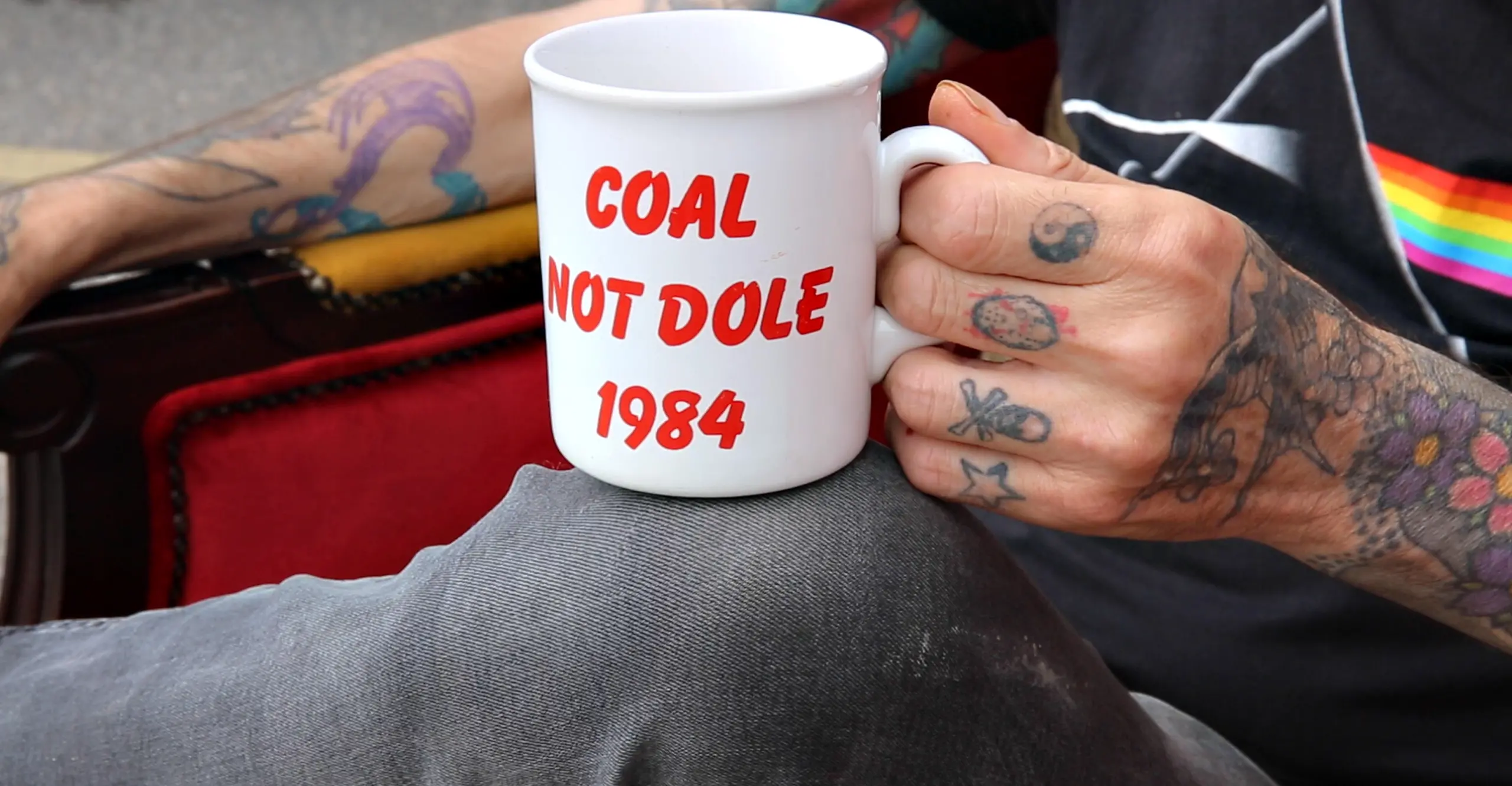 A tattooed hand holds a cup that reads 'Coal Not Dole' 