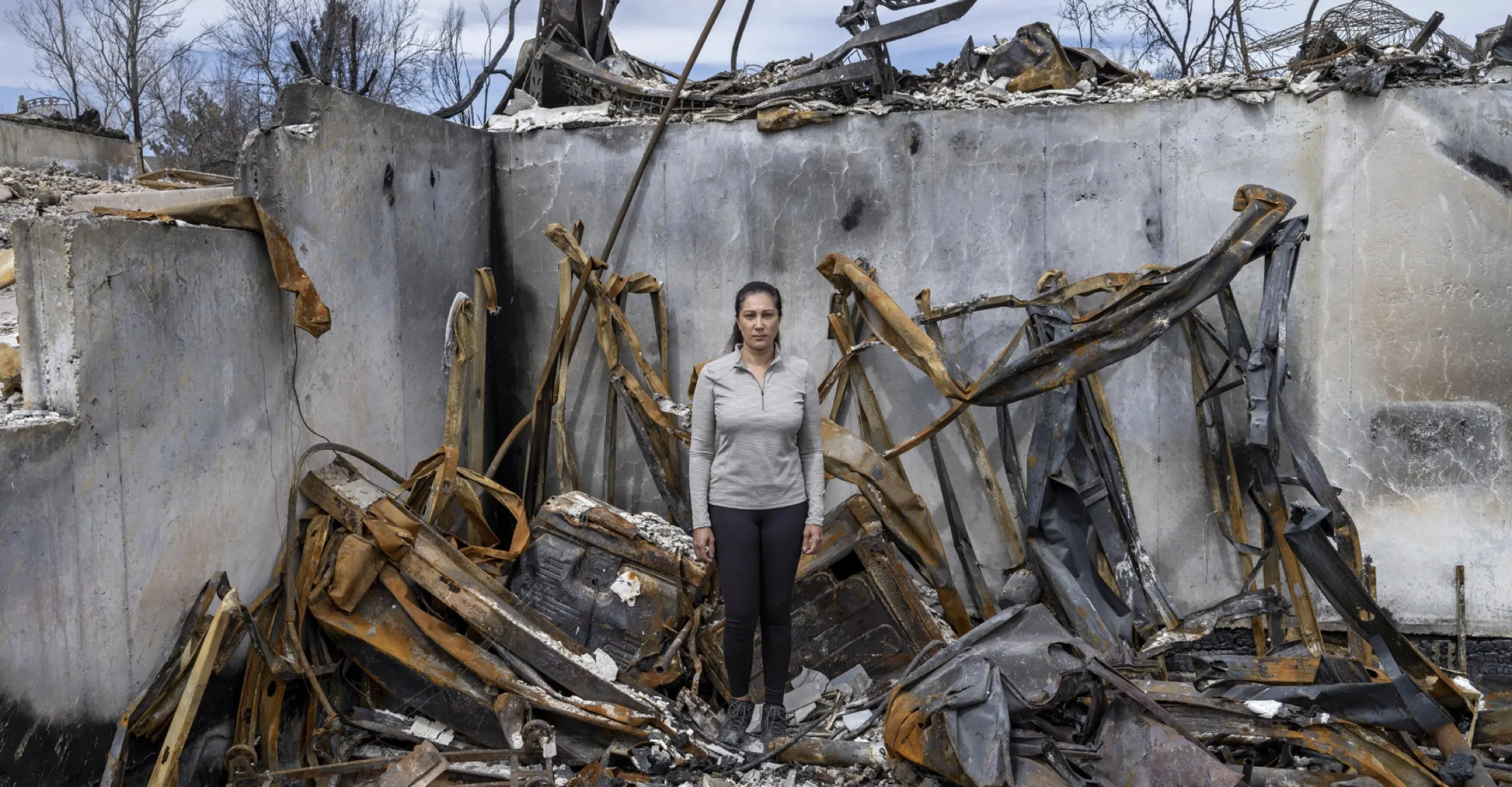 Colour photograph featuring a female presenting person standing in the ruins of a home destroyed by fire
