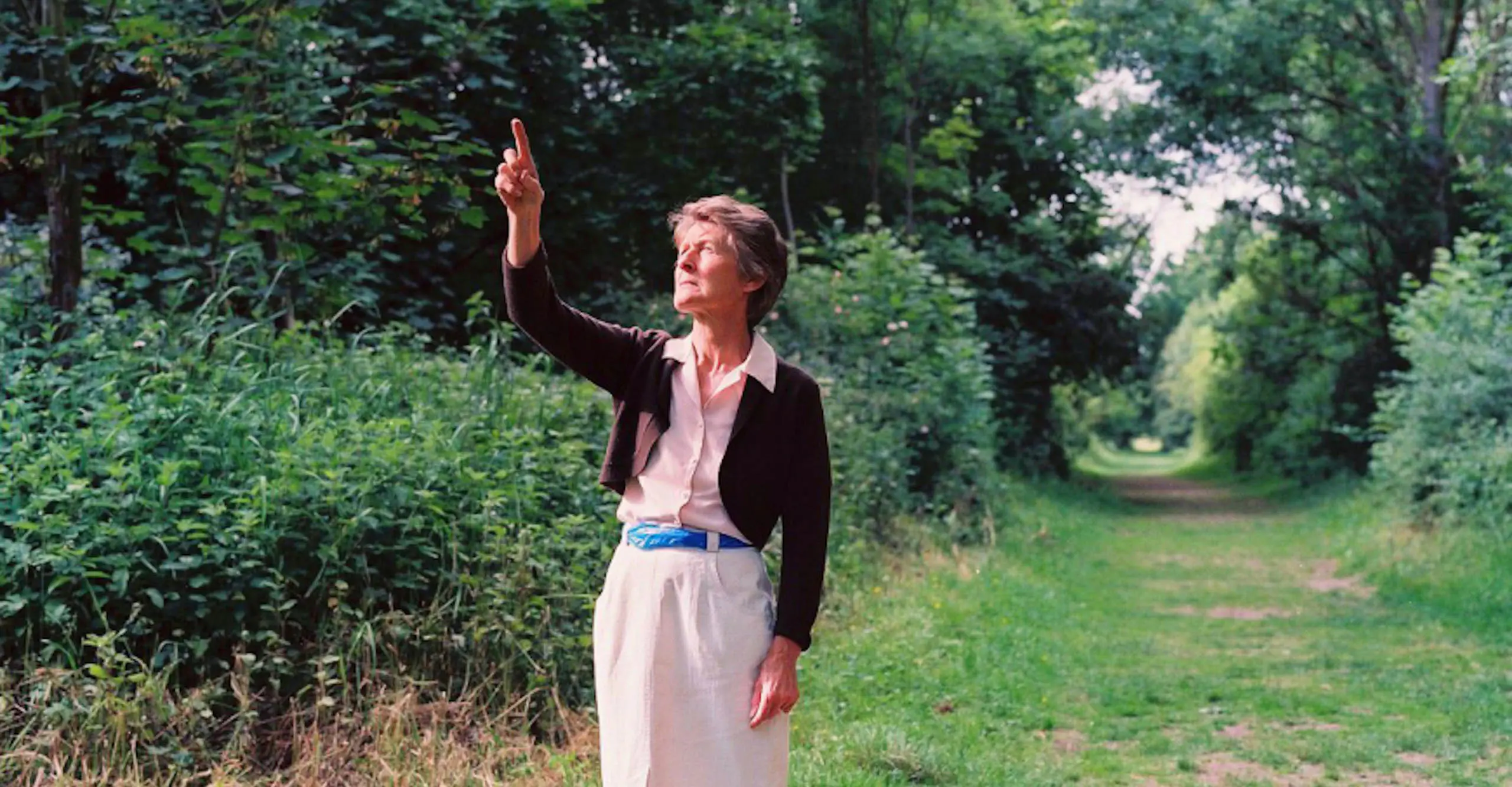 A woman stands next to a field pointing skyward. Around her waist like a belt is a blue plastic bag.