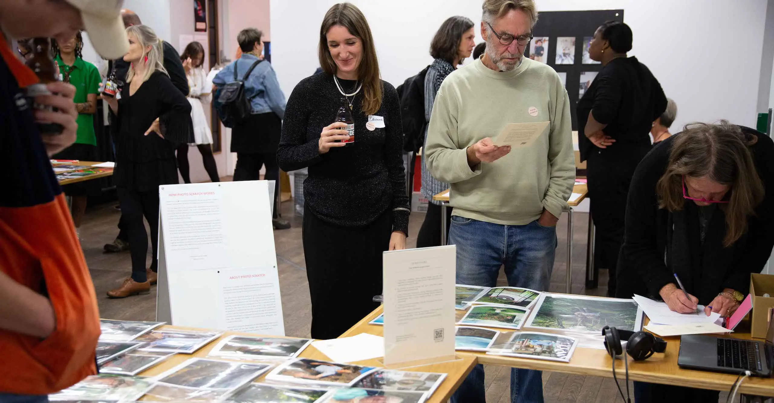 Photo of small groups of people standing around different tables with photographs on them.