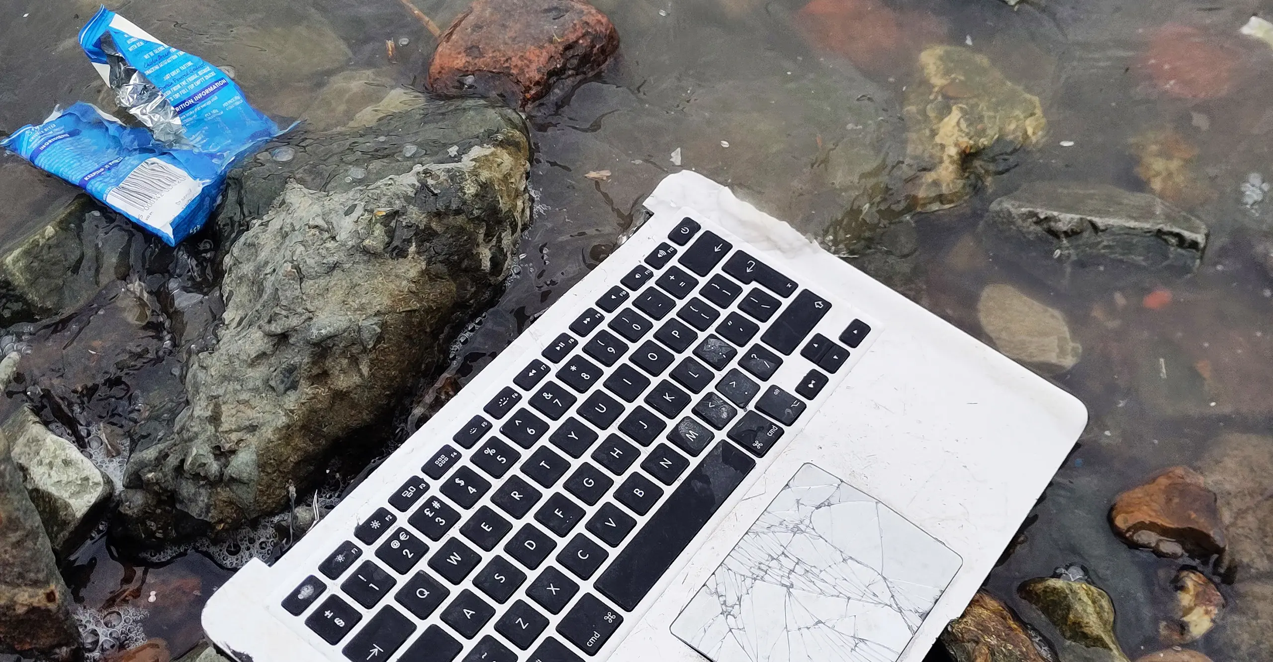 A broken laptop keyboard and an empty bag of crisps sitting ontop of a rocky foreshore.