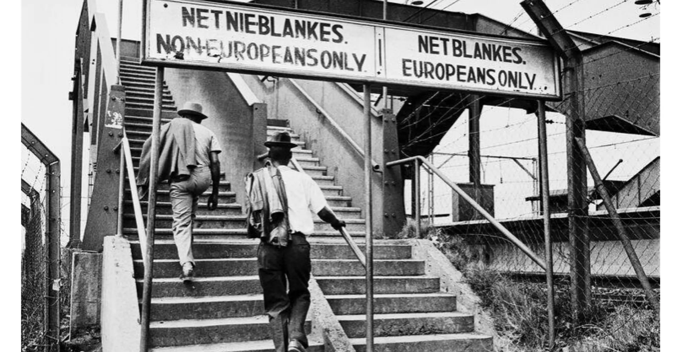 Black and white photograph of a railway bridge staircase with two signs, one saying "Non-Europeans Only" and "Europeans Only". Two black men walk up the "Non-Europeans Only" staircase.