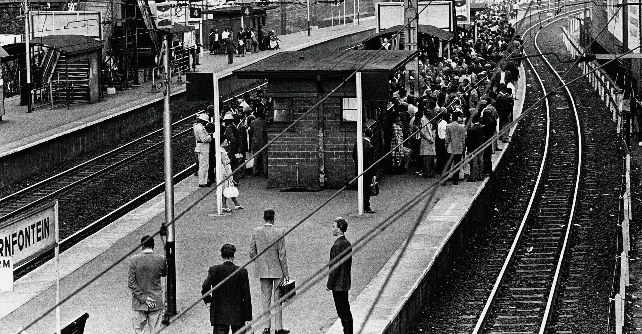 Black and white photograph showing a railway station platform in rush hour with crowds of Black people in the background tightly cramped together while a few white people stand on the almost empty platform, showing the reality of apartheid