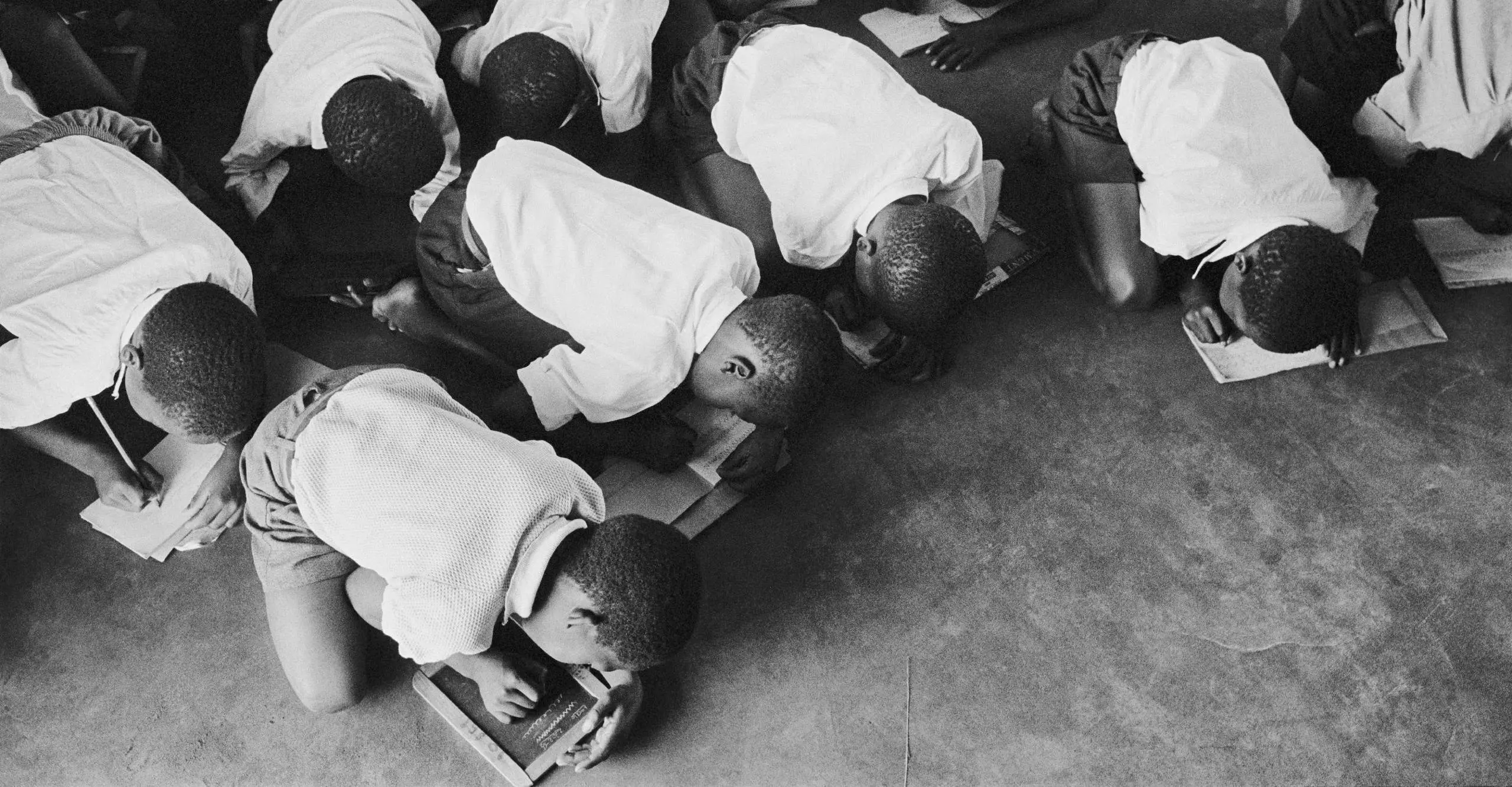 Black and white photograph of several schoolchildren bent over on the floor to write, some write on paper others write on chalkboards.