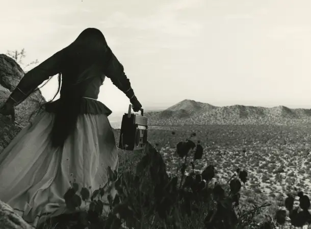 Black and white image of a woman walking in the desert, her back is to the camera as she walks away