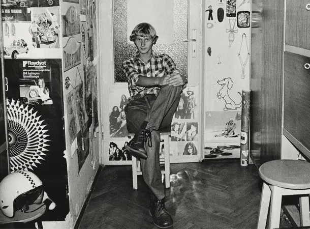 Black and white image of a young person sat inside a domestic room turned toward the camera.
