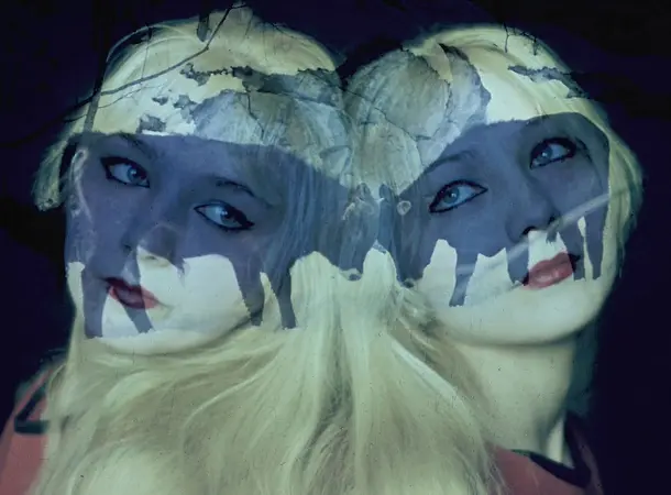 A double exposed photograph of two blonde women with their backs against one another, looking forward toward the camera. An image of two cows with their noses touching is overlayed over their faces.