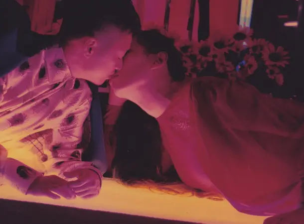 A darkened bar lit with a pink glow. A couple lean across it to meet in a kiss.