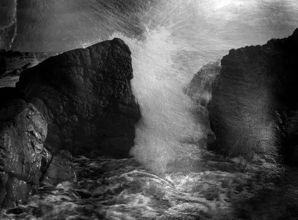 Black and white photograph of ocean waves crashing against large, jagged rocks along a rocky shoreline.