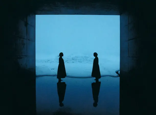 Colour Photograph of two silhouetted figures standing opposite each other inside a dark tunnel with a blue toned expanse of snow in the background. Reflections of the figures appear on the water-covered ground.