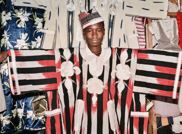 Colour photograph of a young man wearing a large traditional shirt. Behind him a number of Burkinabe textiles can be seen hanging