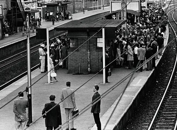 Black and white photograph showing a railway station platform in rush hour with crowds of Black people in the background tightly cramped together while a few white people stand on the almost empty platform, showing the reality of apartheid