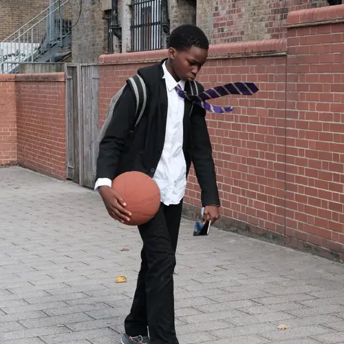 Colour photograph of a young person wearing a school uniform walking, holding a basketball and looking down at the path