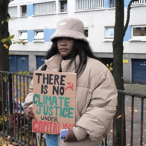 Colour photograph of a young person stood looking at the camera, holding a cardboard sign with the words There is no climate justice under capitalism