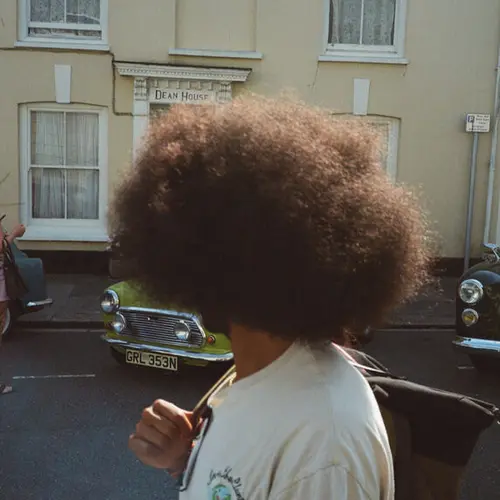 Colour image of person with their head turned obscuring their face, stood in a residential street with a car in the background