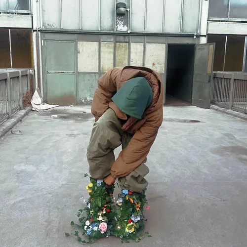 A man stood in front of a block of flats, leaning down to touch his boots whcih are made of foliage and flowers.